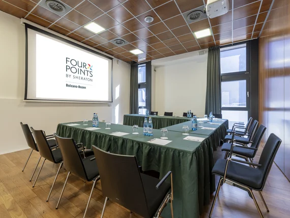 Sala riunioni con tavoli a U, sedie nere e schermo proiettore Four Points Bolzano
